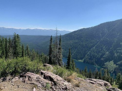 A scenic view of a lush green valley with a lake, surrounded by mountains under a clear blue sky.