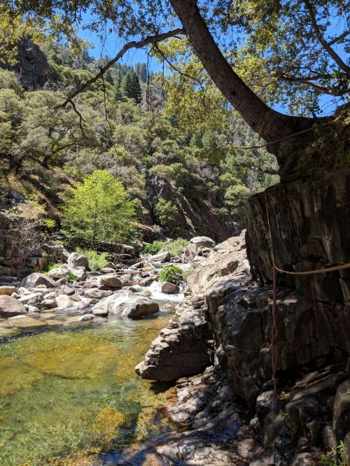 A serene river flows through rocky terrain, surrounded by lush greenery and steep hills under a clear blue sky.