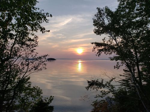 A serene sunset over calm waters, framed by trees on either side, with soft clouds in the sky.