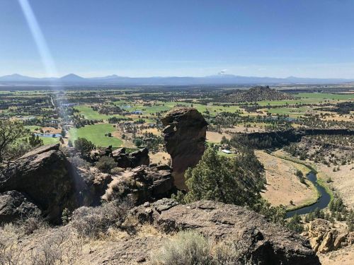 A panoramic view of a valley with green fields, a river, and distant mountains under a clear blue sky.