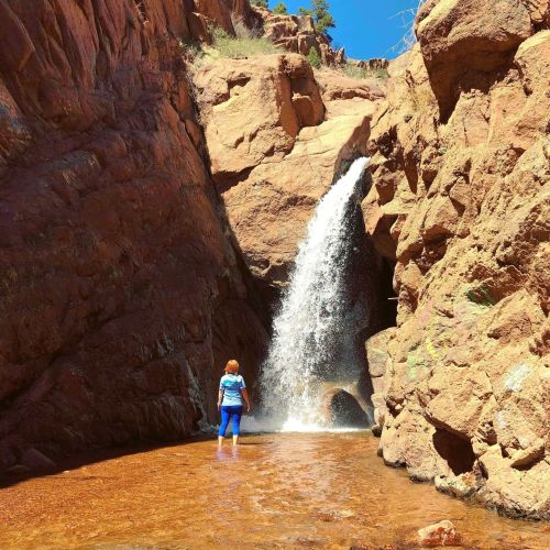 A person stands in shallow water near a waterfall, surrounded by rocky cliffs and greenery.