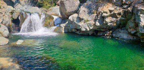 A serene natural pool with clear green water, surrounded by rocks and a small waterfall cascading into it.