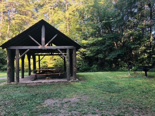 A wooden picnic shelter with tables, surrounded by trees and a grassy area, with a grill nearby.