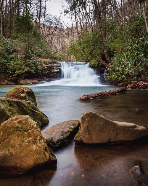 A serene waterfall cascades over rocks into a clear stream, surrounded by lush greenery and trees.