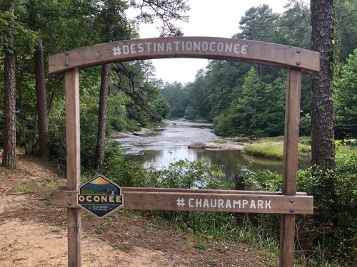 A wooden sign frames a serene river scene in a forested area, promoting Oconee and Chauram Park.
