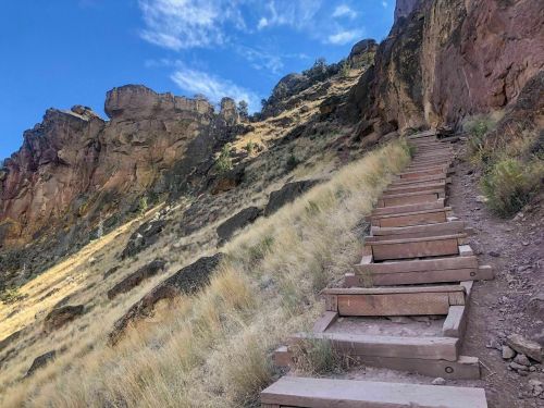 A wooden staircase leads up a grassy hillside, surrounded by rocky cliffs under a blue sky with scattered clouds.