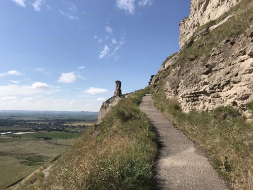 A winding path along a rocky cliff with green grass and a clear blue sky in the background.