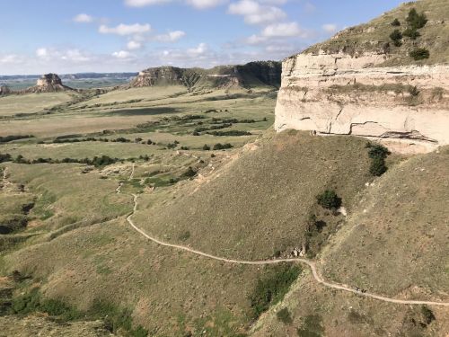 A scenic view of rolling green hills and a winding path, with rocky cliffs under a partly cloudy sky.