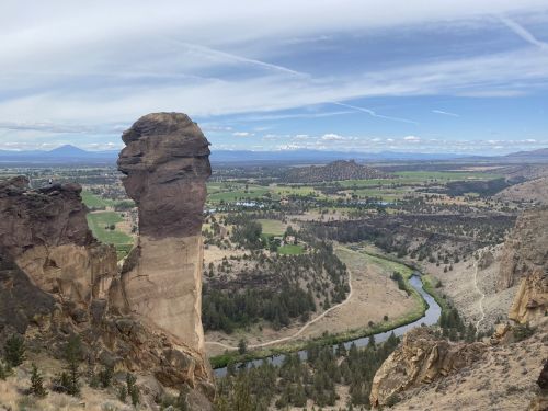 A towering rock formation overlooks a lush valley with a winding river and distant mountains under a cloudy sky.