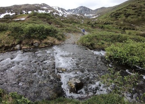 A cyclist rides along a rocky path beside a flowing stream in a mountainous landscape with snow-capped peaks.