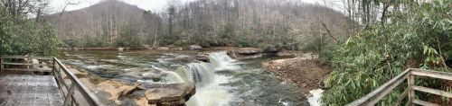 A panoramic view of a river with waterfalls, surrounded by trees and a misty, overcast sky.