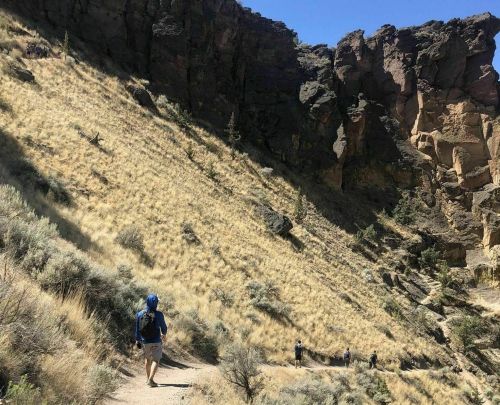 A hiker walks along a trail in a rocky, sunlit landscape with dry grass and steep cliffs in the background.