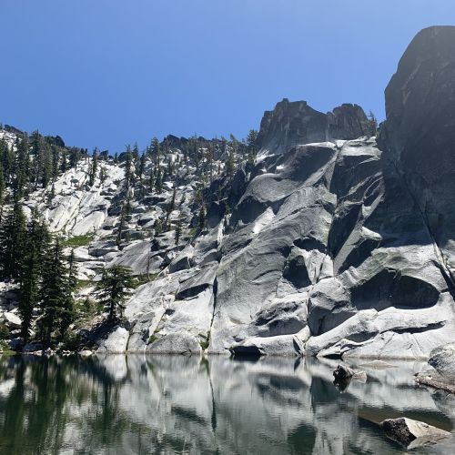 A serene lake reflects towering granite cliffs and lush trees under a clear blue sky.