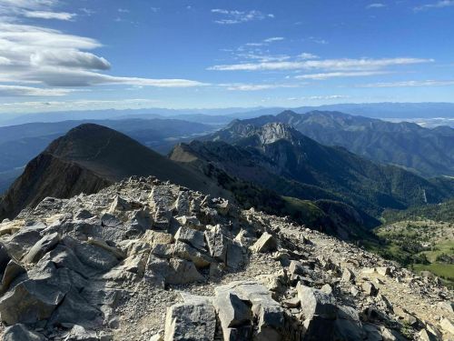 A panoramic view from a mountain summit, showcasing rocky terrain and distant green hills under a blue sky.