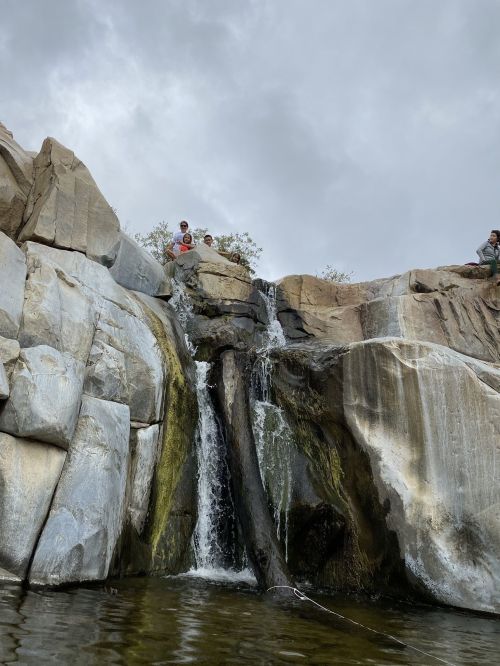 A small waterfall cascades over rocky cliffs, with two people standing at the top, under a cloudy sky.