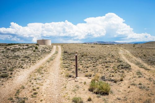 A dirt road splits in a barren landscape, leading to a water tank under a blue sky with fluffy clouds.