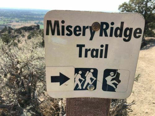 Sign for Misery Ridge Trail with directional arrow and hiker icons, set against a scenic landscape.