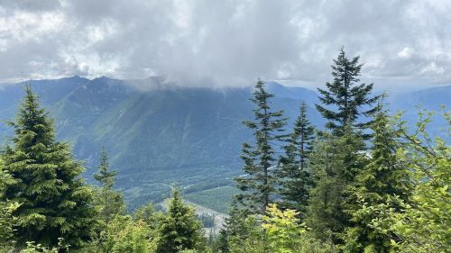 A scenic view of mountains shrouded in clouds, surrounded by lush green trees under a partly cloudy sky.