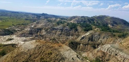 A panoramic view of rugged hills and valleys under a blue sky, showcasing a mix of greenery and rocky terrain.