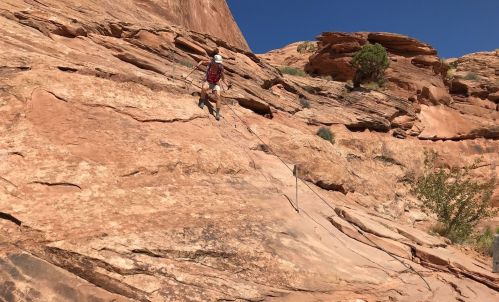 A climber ascends a rocky cliff with ropes, surrounded by red rock formations and clear blue skies.