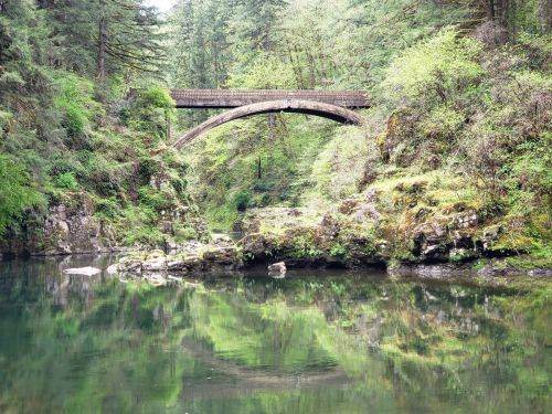 A stone bridge arches over a serene river, surrounded by lush green trees and rocky banks reflecting in the water.