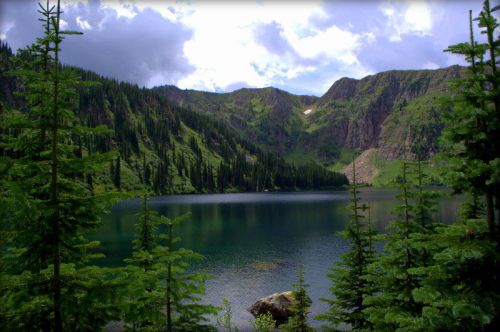 A serene lake surrounded by lush green mountains and pine trees under a cloudy sky.