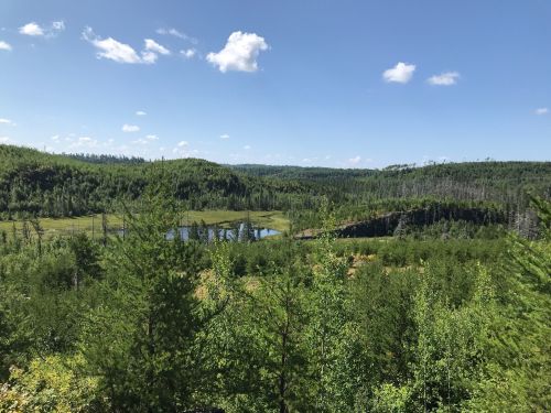 Lush green landscape with rolling hills, a river, and a clear blue sky dotted with clouds.