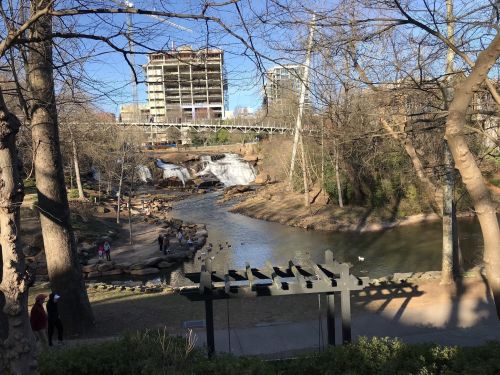 A scenic view of a river with a waterfall, surrounded by trees and a city skyline in the background.