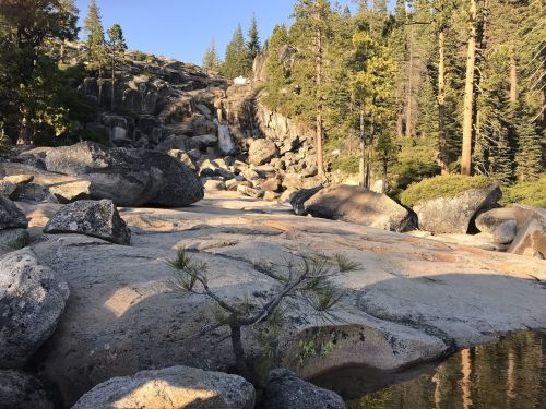 A serene landscape featuring large boulders, a gentle waterfall, and tall trees under a clear blue sky.
