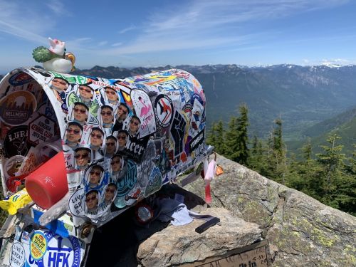 A decorated mailbox covered in stickers sits on a rocky outcrop, overlooking a scenic mountain landscape.