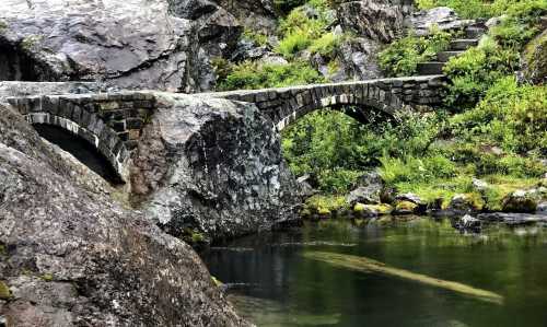 A stone arch bridge over a calm, reflective pond surrounded by lush greenery and rocky terrain.