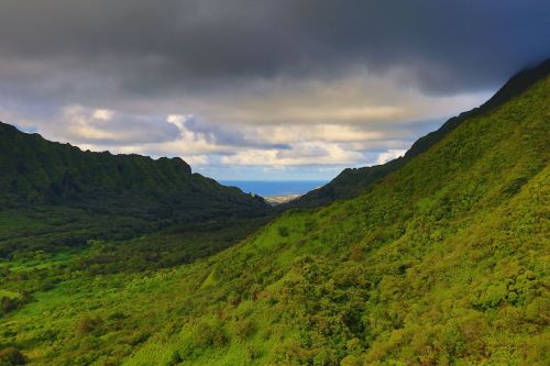 Lush green mountains frame a valley leading to the ocean under a cloudy sky.