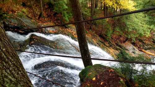 A rushing waterfall flows over rocky terrain, surrounded by trees and autumn leaves.
