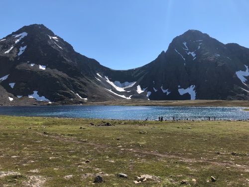 A serene mountain landscape featuring two peaks, a clear blue lake, and patches of snow under a bright blue sky.