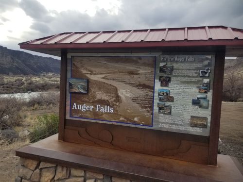 Sign for Auger Falls with historical information, set against a backdrop of a river and rocky landscape.