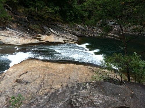 A serene river flows over smooth rocks, surrounded by lush greenery and steep, rocky banks.