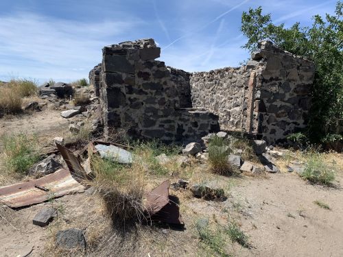 A partially collapsed stone structure surrounded by dry grass and debris under a clear blue sky.