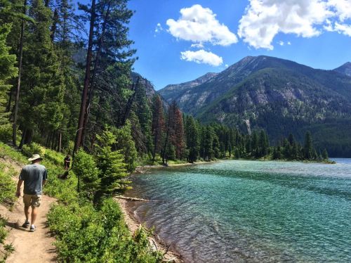 Two hikers walk along a scenic trail by a clear lake, surrounded by lush trees and mountains under a bright blue sky.