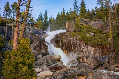 A waterfall cascades over rocky terrain, surrounded by lush green trees and a clear blue sky.