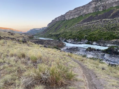 A winding river flows through a rocky valley surrounded by green hills under a clear sky at sunset.