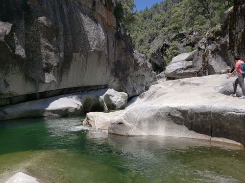 A person walks along a rocky riverbank beside clear green water, surrounded by steep, rugged cliffs and trees.
