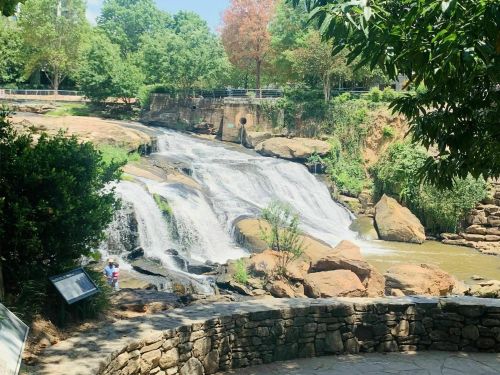 A scenic waterfall surrounded by greenery, with a small child playing nearby and a stone wall in the foreground.