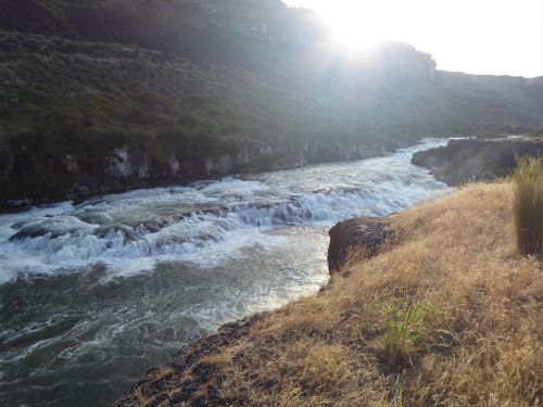 A river flows over rocks, surrounded by grassy banks and hills, with the sun shining in the background.