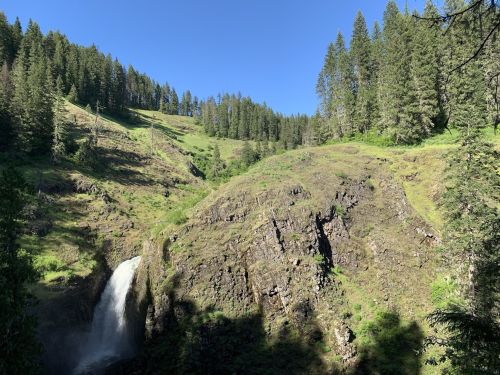 A scenic view of a waterfall cascading down rocky terrain, surrounded by lush green hills and tall pine trees under a clear blue sky.