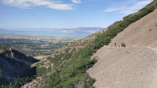 Hikers on a winding trail along a mountainside, overlooking a valley and lake under a clear blue sky.