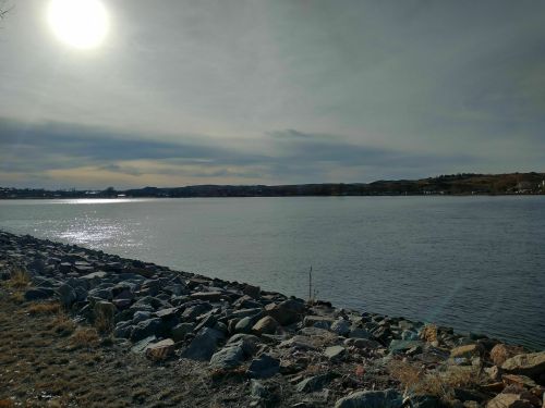 A serene river scene with rocky shorelines under a cloudy sky and a bright sun reflecting on the water.