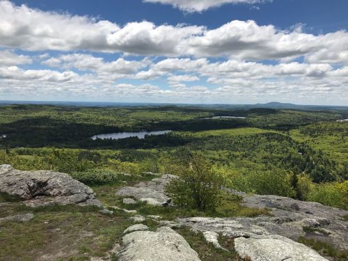 A panoramic view of lush green hills and a lake under a bright blue sky with fluffy white clouds.