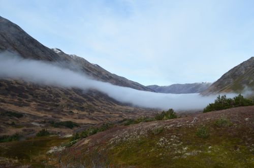 A serene valley surrounded by mountains, with a layer of fog drifting through the landscape under a clear sky.