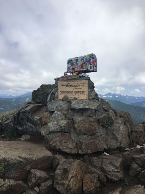 A colorful, decorated box sits atop a rocky outcrop with a plaque, surrounded by mountains and cloudy skies.