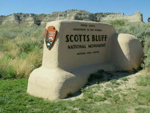 Sign for Scotts Bluff National Monument, featuring the National Park Service logo, surrounded by grass and rocky cliffs.